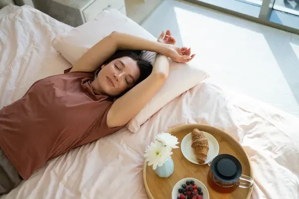 Femme allongée paisiblement dans son lit, avec un petit-déjeuner au réveil, symbolisant l’importance du bien-être pour une vie sexuelle épanouie.